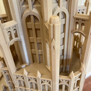 May include: A detailed 3D printed model of a tall building with intricate architectural details, including pointed arches, windows, and a spire. The model is made of a light beige plastic and appears to be a replica of a historic building.