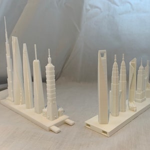 May include: Two sets of white miniature skyscraper models. Each set is mounted on a rectangular base. The buildings vary in design, including towers with spires, rounded tops, and rectangular structures. The models are likely 3D printed.