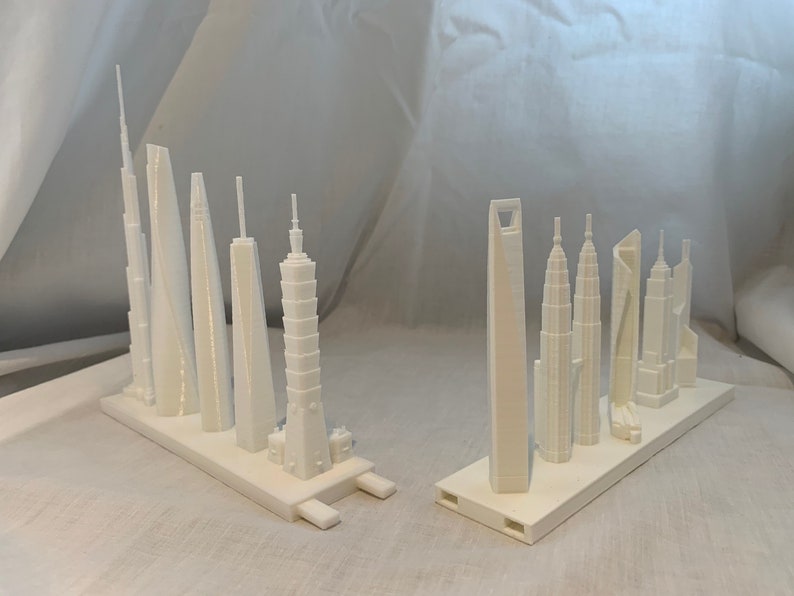 May include: Two sets of white miniature skyscraper models. Each set is mounted on a rectangular base. The buildings vary in design, including towers with spires, rounded tops, and rectangular structures. The models are likely 3D printed.