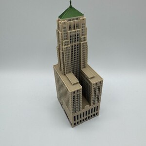 May include: A miniature beige skyscraper model with a green roof. The building has many windows and architectural details. The model is a detailed representation of a tall building.