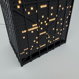 May include: A black, geometric, illuminated lamp shaped like a skyscraper. The lamp has many small, square, yellow lights that resemble windows. The lamp is on a white surface.