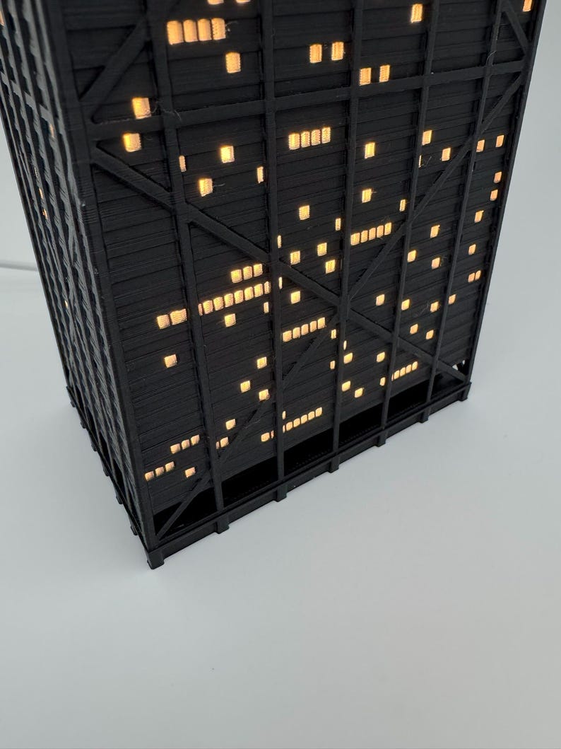 May include: A black, geometric, illuminated lamp shaped like a skyscraper. The lamp has many small, square, yellow lights that resemble windows. The lamp is on a white surface.