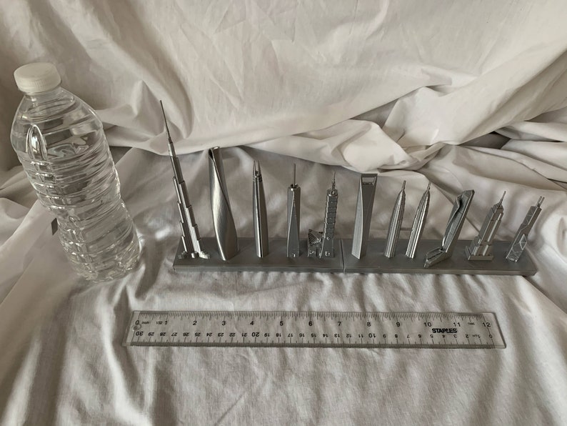 May include: A collection of silver-colored miniature skyscraper models displayed on a gray base. A clear plastic water bottle and a ruler are also in the image. The ruler measures up to 12 inches.