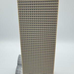 May include: A beige and gray architectural model of a skyscraper. The building has a grid-like facade with numerous small square windows. The model is set on a rectangular base, with a smaller structure attached to the main tower.