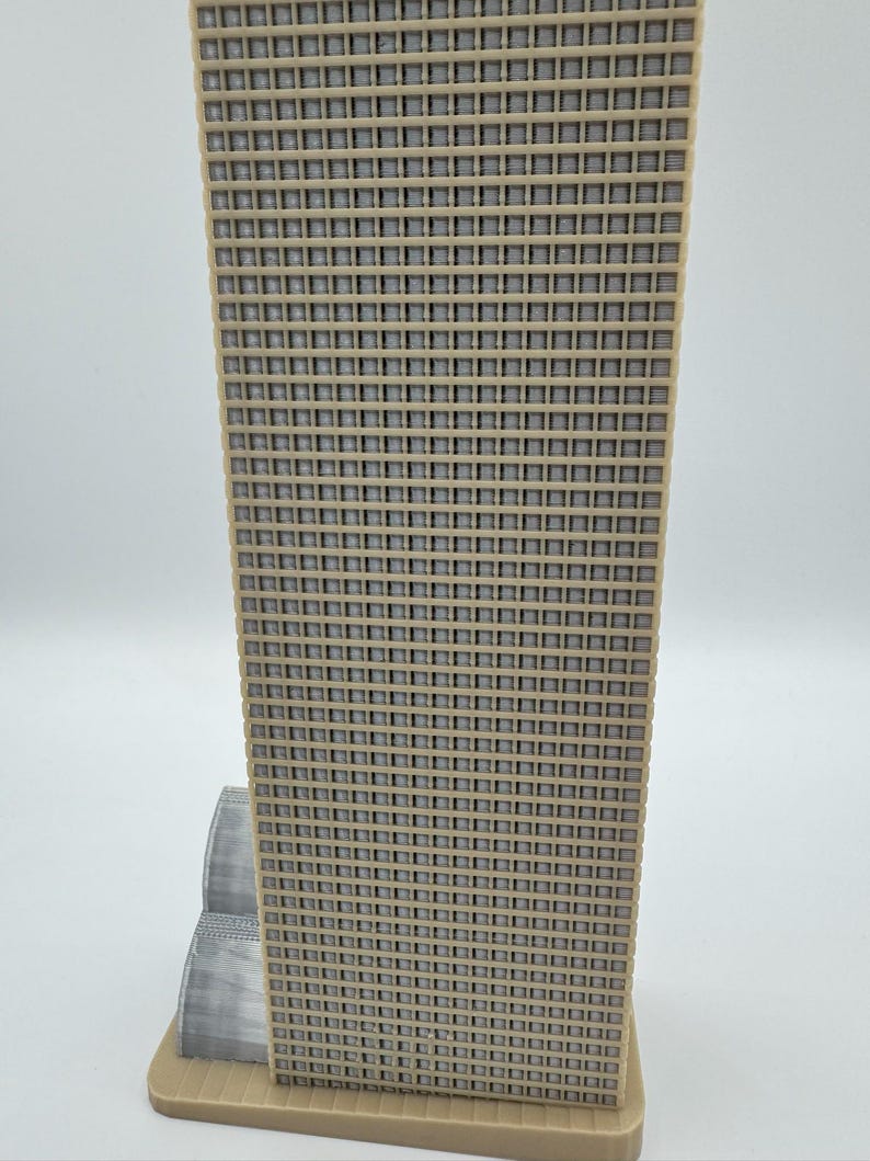 May include: A beige and gray architectural model of a skyscraper. The building has a grid-like facade with numerous small square windows. The model is set on a rectangular base, with a smaller structure attached to the main tower.