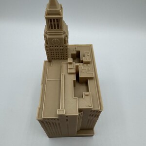 May include: A tan-colored architectural model of a building with a clock tower. The model has detailed features, including windows and roof structures. The clock tower has a clock face. The model is likely 6 inches tall.