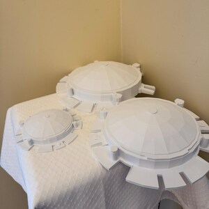 May include: Three white, dome-shaped objects with circular bases and geometric designs are arranged on a white, textured tablecloth. The objects vary in size, with the largest in the foreground and the smallest in the background.