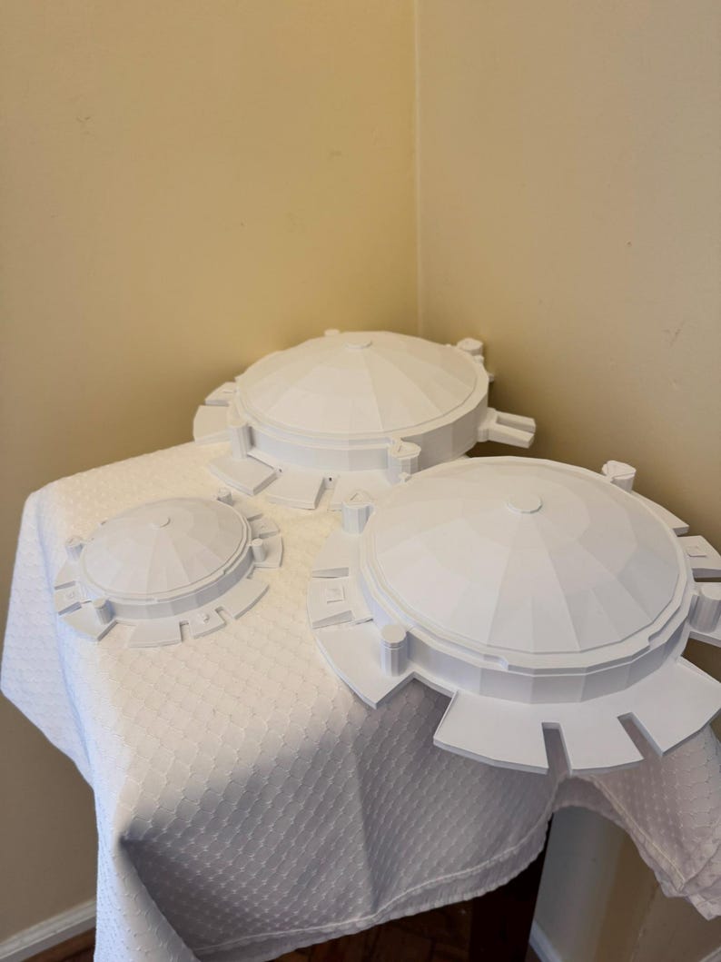 May include: Three white, dome-shaped objects with circular bases and geometric designs are arranged on a white, textured tablecloth. The objects vary in size, with the largest in the foreground and the smallest in the background.