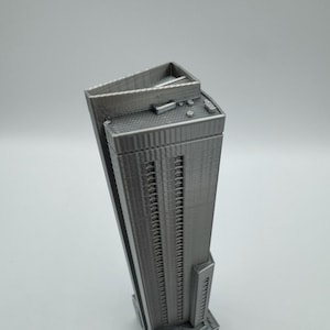 May include: A gray, 3D-printed model of a skyscraper. The building has a rectangular shape with many windows and a flat roof. The model is detailed, showing the texture of the building's facade and roof.
