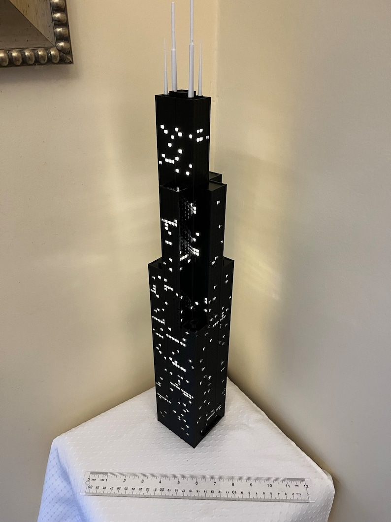 May include: A black and white model of a skyscraper with white antenna on top. The model is sitting on a white surface with a ruler in the foreground.