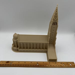 May include: A beige architectural model of a church with a tall steeple and a long, rectangular main structure. The model is placed on a white surface with a wooden ruler nearby, showing measurements in inches.