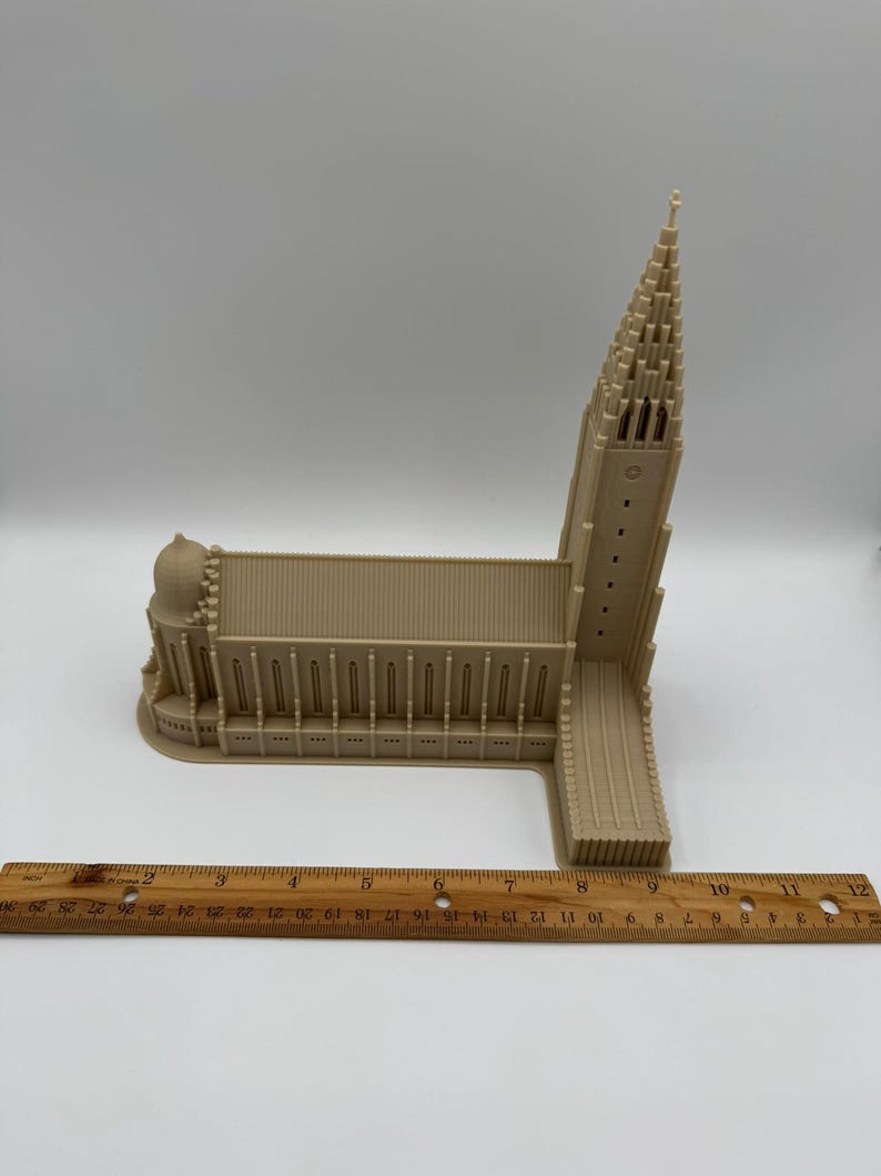 May include: A beige architectural model of a church with a tall steeple and a long, rectangular main structure. The model is placed on a white surface with a wooden ruler nearby, showing measurements in inches.