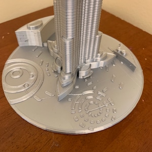 May include: A silver 3D printed model of a skyscraper with a detailed base.