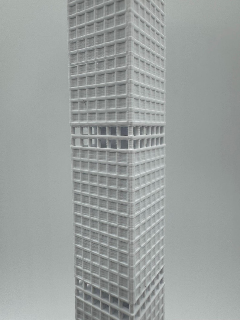 May include: A white, 3D-printed model of a skyscraper. The rectangular building features a grid of windows and open sections. The model is a detailed architectural representation, likely made using a 3D printer.