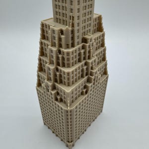 May include: A beige, 3D-printed model of a skyscraper. The building has a stepped design with numerous windows and architectural details. The model is a detailed representation of a classic building.