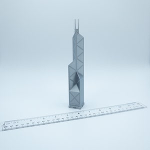 May include: A gray 3D-printed model of a tall building with a unique architectural design. The model is placed next to a clear ruler, providing a size reference. The building has a geometric facade and two vertical spires at the top.