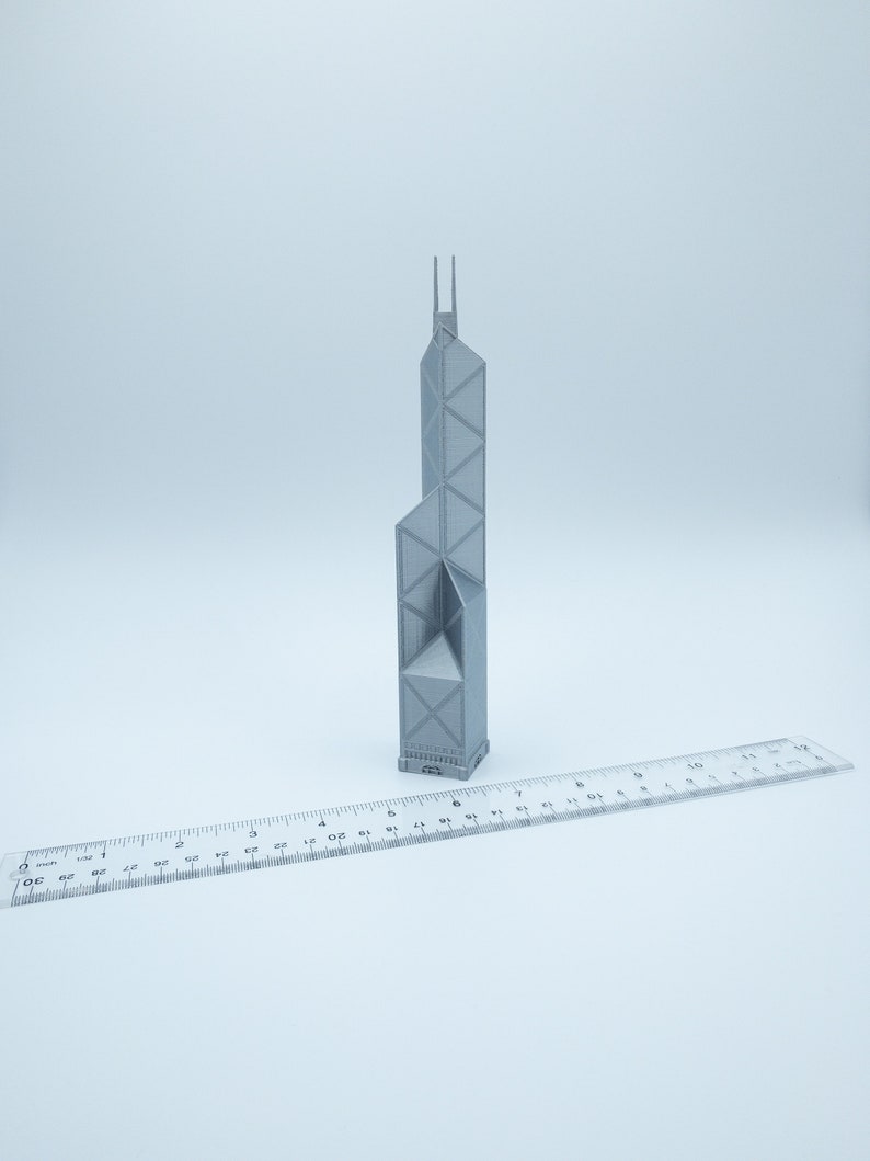 May include: A gray 3D-printed model of a tall building with a unique architectural design. The model is placed next to a clear ruler, providing a size reference. The building has a geometric facade and two vertical spires at the top.