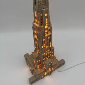 May include: A beige, 3D-printed architectural model of the Cathedral of Learning, featuring a tall, multi-tiered tower with intricate gothic-style window cutouts. The structure is illuminated from within, casting a warm, orange glow through the windows.