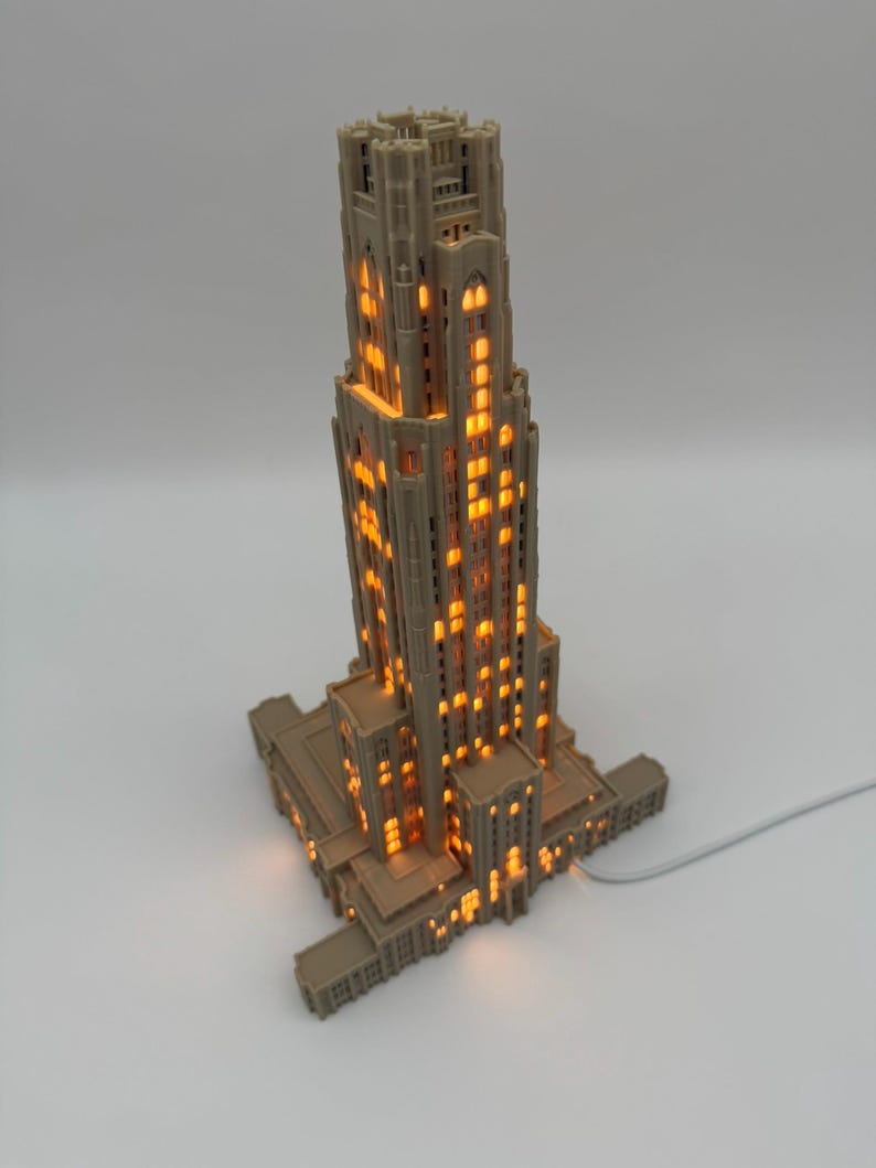 May include: A beige, 3D-printed architectural model of the Cathedral of Learning, featuring a tall, multi-tiered tower with intricate gothic-style window cutouts. The structure is illuminated from within, casting a warm, orange glow through the windows.