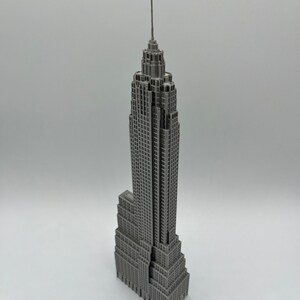 K&ouml;nnte beinhalten: Ein graues Miniaturmodell des Empire State Buildings. Das Modell ist eine detaillierte Nachbildung des ikonischen Wolkenkratzers mit mehreren Etagen und einer hohen Antenne. Das Geb&auml;ude ist grau und steht auf einer wei&szlig;en Oberfl&auml;che.