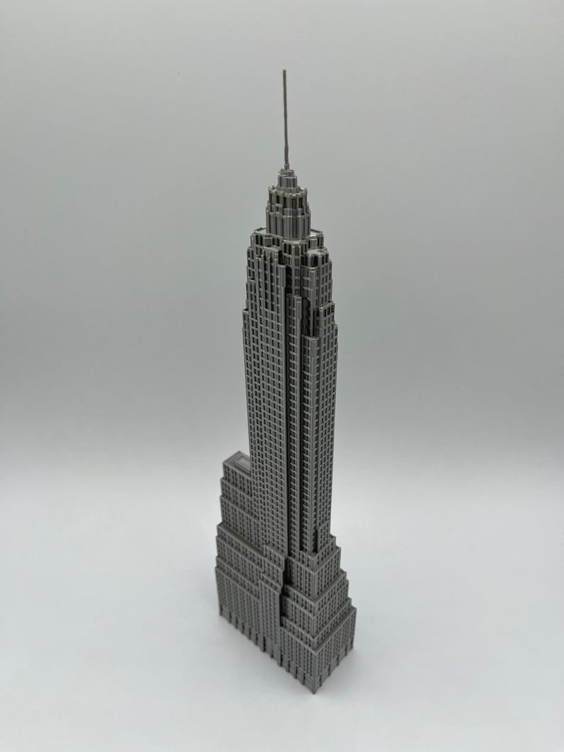 K&ouml;nnte beinhalten: Ein graues Miniaturmodell des Empire State Buildings. Das Modell ist eine detaillierte Nachbildung des ikonischen Wolkenkratzers mit mehreren Etagen und einer hohen Antenne. Das Geb&auml;ude ist grau und steht auf einer wei&szlig;en Oberfl&auml;che.