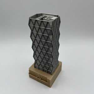 May include: A 3D printed model of a modern skyscraper with a geometric facade. The building is made of light grey plastic and has a base made of light brown plastic.