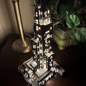 May include: A miniature black skyscraper model featuring intricate architectural details and multiple illuminated windows that emit a warm white glow, creating a realistic night-time city building effect with a tiered structure and a rectangular base.