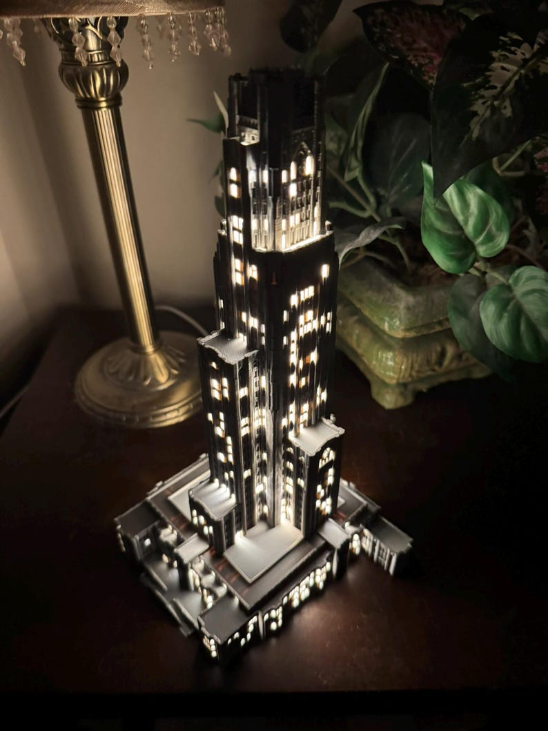 May include: A miniature black skyscraper model featuring intricate architectural details and multiple illuminated windows that emit a warm white glow, creating a realistic night-time city building effect with a tiered structure and a rectangular base.