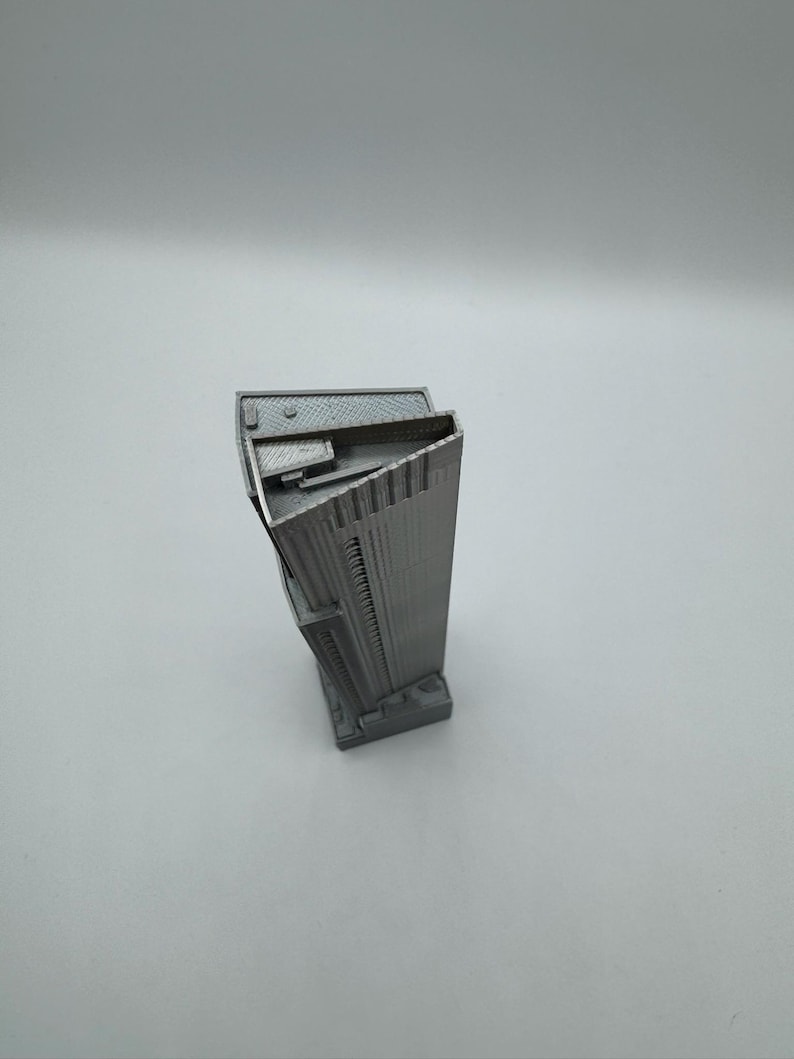 May include: A gray, 3D-printed miniature skyscraper model. The building has a rectangular base and a tall, multi-story design with detailed architectural features. The model is made of a solid, matte gray material.