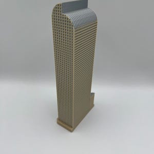 May include: A tan and gray architectural model of a skyscraper. The building has a rectangular shape with a grid-like facade and a curved top. The model is made of plastic and sits on a white surface.