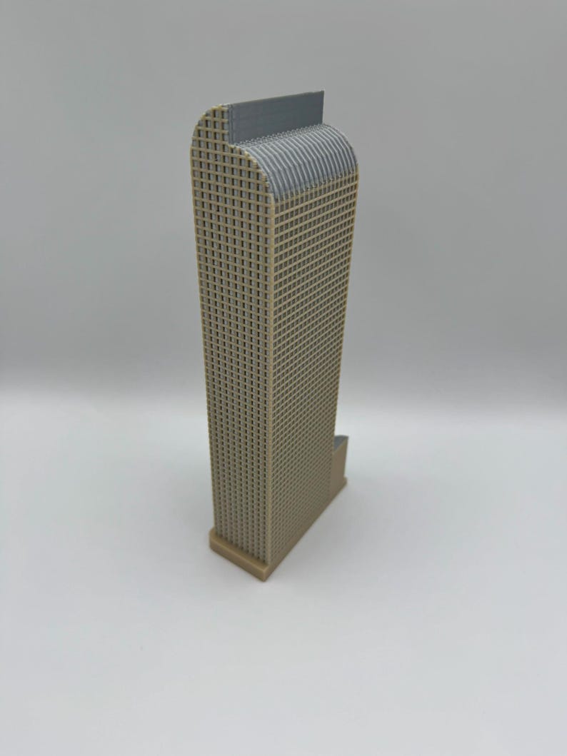 May include: A tan and gray architectural model of a skyscraper. The building has a rectangular shape with a grid-like facade and a curved top. The model is made of plastic and sits on a white surface.
