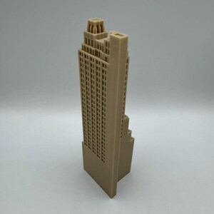 May include: A tan-colored, 3D-printed model of a skyscraper. The building features a detailed facade with numerous windows and a stepped design, typical of Art Deco architecture. The model stands on a rectangular base.