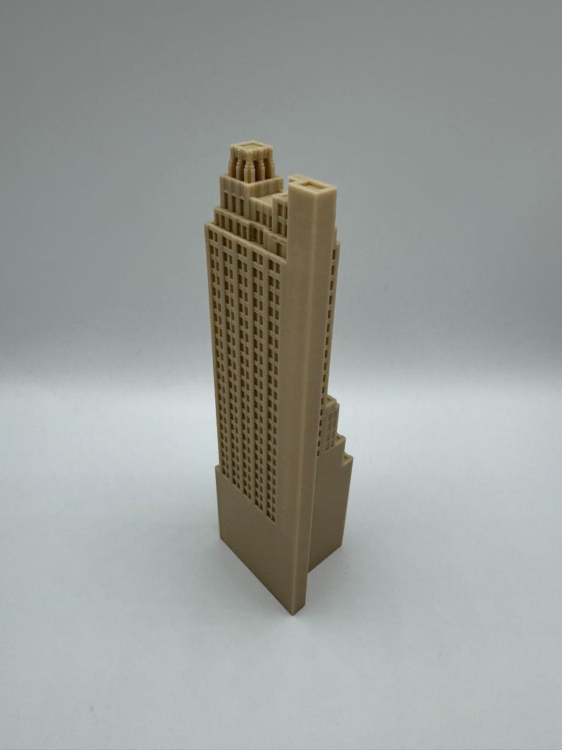 May include: A tan-colored, 3D-printed model of a skyscraper. The building features a detailed facade with numerous windows and a stepped design, typical of Art Deco architecture. The model stands on a rectangular base.