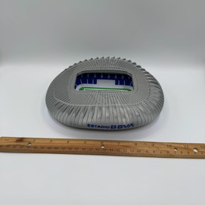 May include: A gray stadium model with a pleated exterior and a green and blue interior. The text "ESTADIO BBVA" is printed in blue on the side. A wooden ruler is placed below the stadium.
