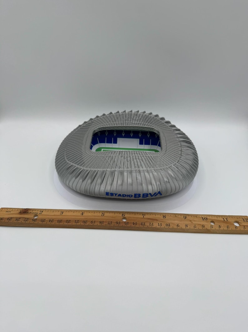 May include: A gray stadium model with a pleated exterior and a green and blue interior. The text "ESTADIO BBVA" is printed in blue on the side. A wooden ruler is placed below the stadium.