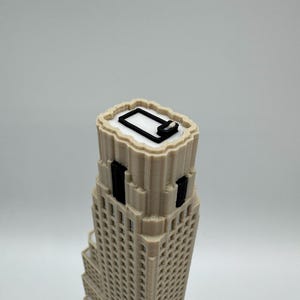 May include: A 3D printed beige skyscraper model featuring a stepped architectural design with vertical black window accents and a white roof surface. The top of the building displays a black rectangular outline and a small white square detail.