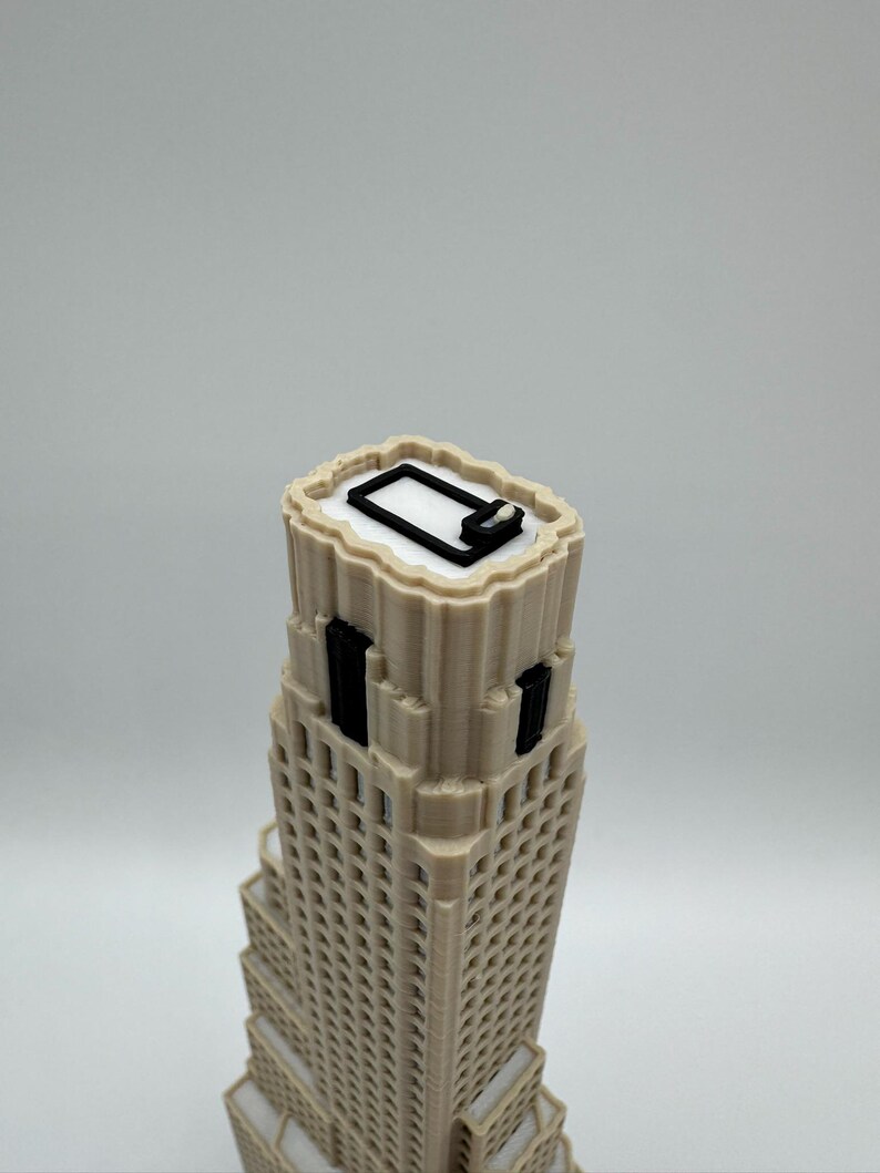 May include: A 3D printed beige skyscraper model featuring a stepped architectural design with vertical black window accents and a white roof surface. The top of the building displays a black rectangular outline and a small white square detail.