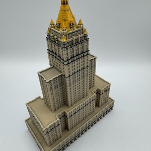 May include: A detailed miniature model of a skyscraper with a beige facade and a gold-colored spire. The building has multiple levels and architectural details, showcasing a classic design. The model is set against a plain white background.