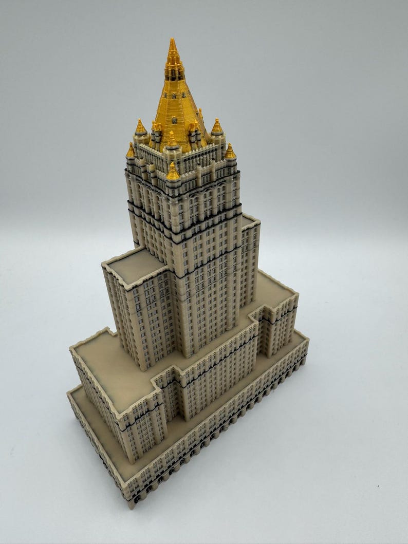 May include: A detailed miniature model of a skyscraper with a beige facade and a gold-colored spire. The building has multiple levels and architectural details, showcasing a classic design. The model is set against a plain white background.