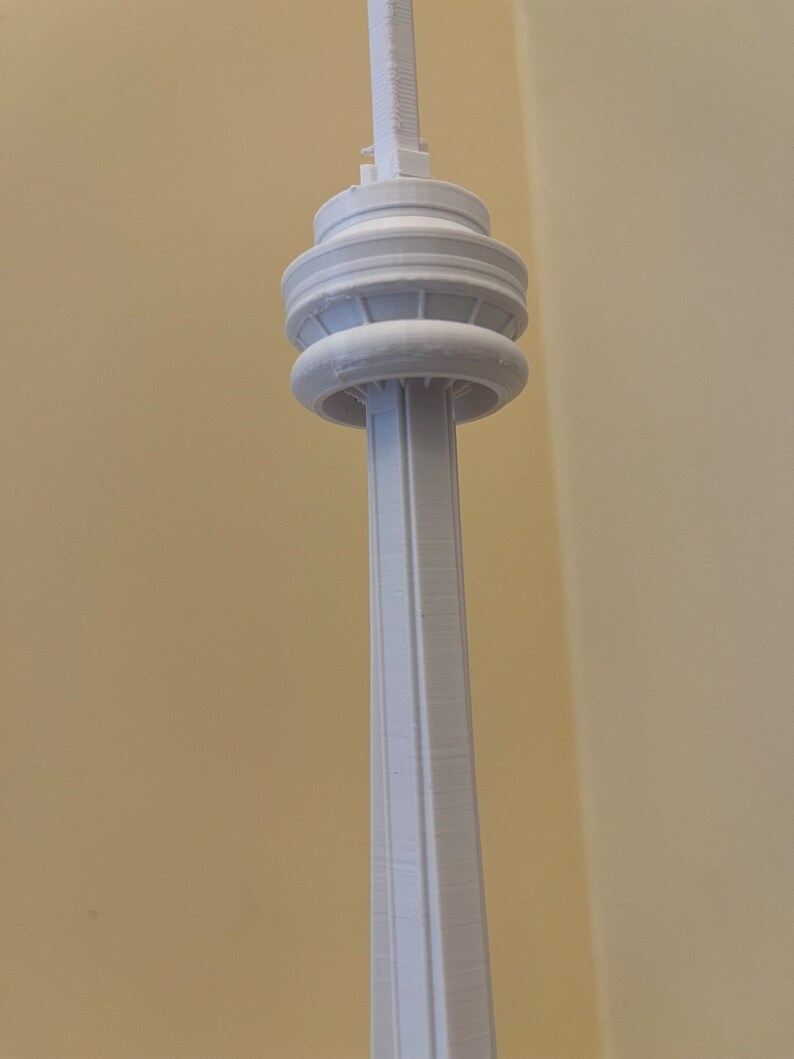May include: A white 3D-printed model of the CN Tower, a prominent landmark in Toronto, Canada. The model features the tower's distinctive cylindrical shaft, observation decks, and antenna. The model is a light, neutral color, and the details are clearly defined.