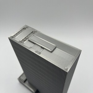 May include: A 3D printed model of a skyscraper with a textured facade. The model is made of a silver-colored material and has a flat roof with a small rectangular opening.