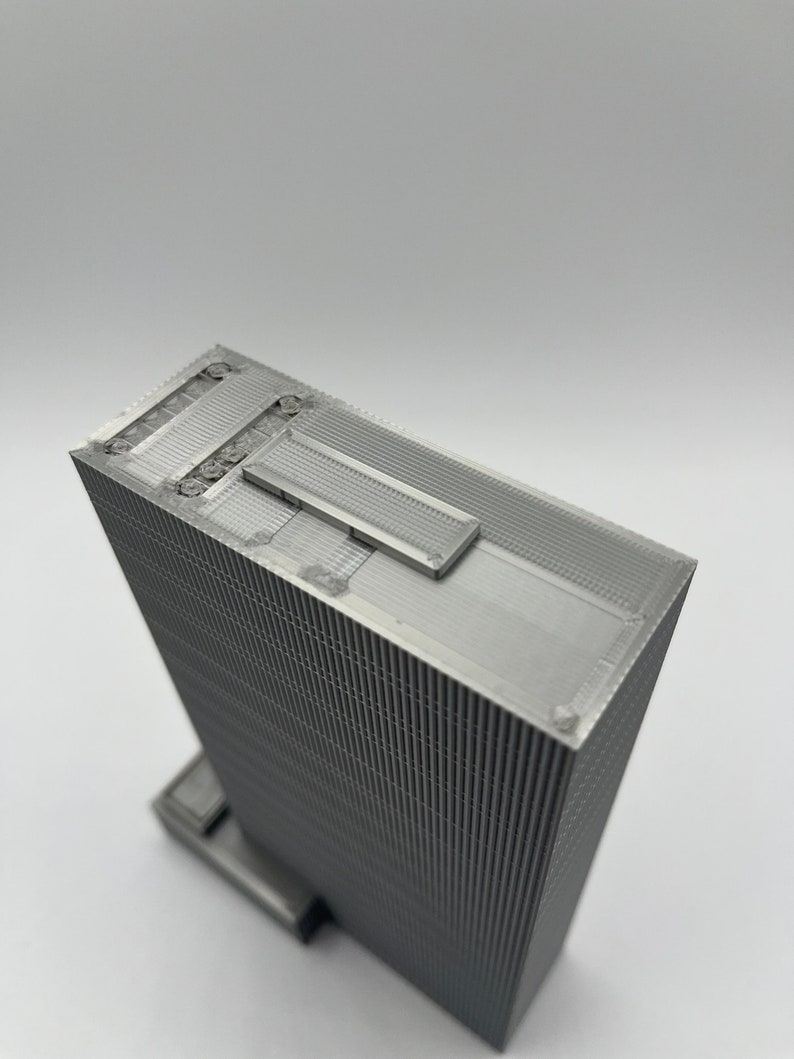 May include: A 3D printed model of a skyscraper with a textured facade. The model is made of a silver-colored material and has a flat roof with a small rectangular opening.