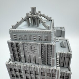 May include: A gray 3D-printed model of a building with the word "EASTERN" embossed on the facade. The model features architectural details, including a clock, decorative elements, and a rooftop structure. The building is a light gray color.