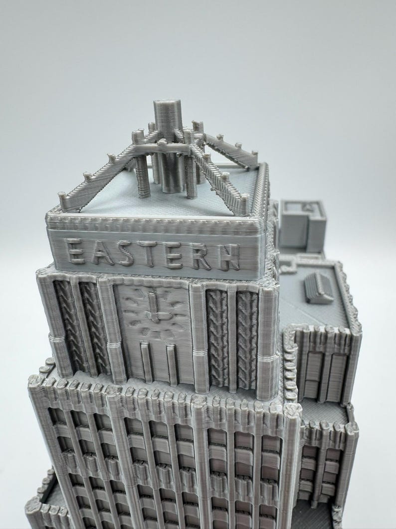 May include: A gray 3D-printed model of a building with the word "EASTERN" embossed on the facade. The model features architectural details, including a clock, decorative elements, and a rooftop structure. The building is a light gray color.