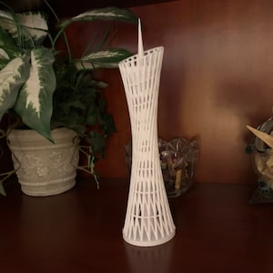 May include: A white 3D printed model of a tall, slender building with a pointed top and a lattice-like design. The model is on a wooden shelf with a potted plant and a bowl of seashells.