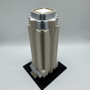 May include: A 3D-printed model of a skyscraper. The building is primarily beige with gray and white accents, and sits on a black square base. The architectural model has a detailed, geometric design.