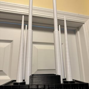 May include: A 3D-printed architectural model with a black base and several white, cylindrical spires of different heights. The model is set against a neutral background, emphasizing the contrast between the dark base and the light spires.