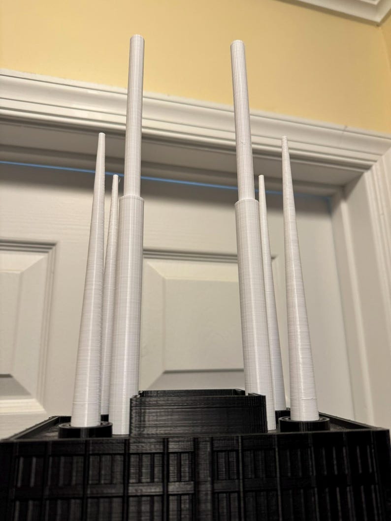 May include: A 3D-printed architectural model with a black base and several white, cylindrical spires of different heights. The model is set against a neutral background, emphasizing the contrast between the dark base and the light spires.