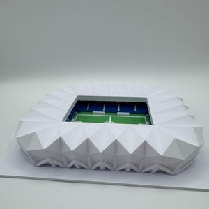 May include: A white, geometric model of a stadium with a green field and blue seating. The stadium has a detailed, angular roof structure and is set on a white base. The model is a miniature representation of a sports venue.