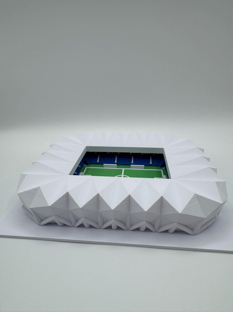 May include: A white, geometric model of a stadium with a green field and blue seating. The stadium has a detailed, angular roof structure and is set on a white base. The model is a miniature representation of a sports venue.